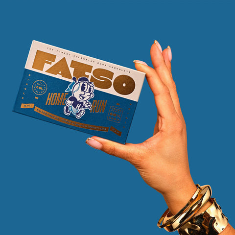 A hand wearing gold bracelets holds a Fatsio Home Run chocolate bar against a solid blue background.