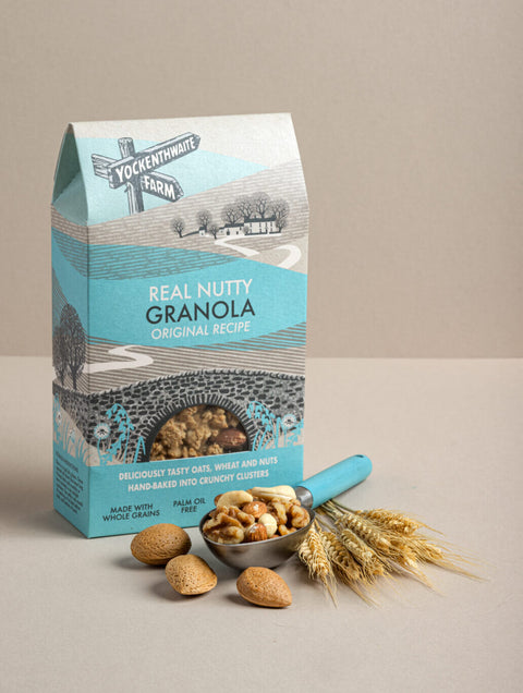 A package of Yockenthwaite Farm Real Nutty Granola Original Recipe stands on a beige surface with a small bowl of mixed nuts, whole almonds, and wheat stalks placed in front. The bag is light blue and white with illustrated graphics, and the bowl contains tan and beige nuts.