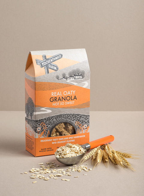 A package of Yockenthwaite Farm Real Oaty Granola (not so sweet) stands on a beige surface with a metal scoop of oats, scattered oats, and wheat stalks in front of it.