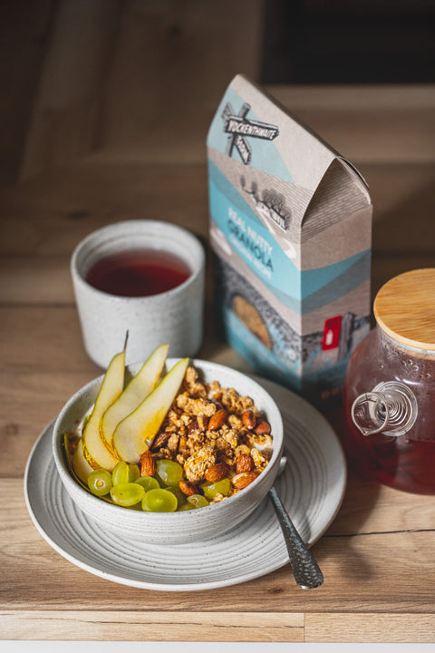 A bowl of granola with sliced pears, green grapes, and nuts on a saucer, with a cup of dark tea, a bag of granola, and a glass jar of tea on a wooden table.