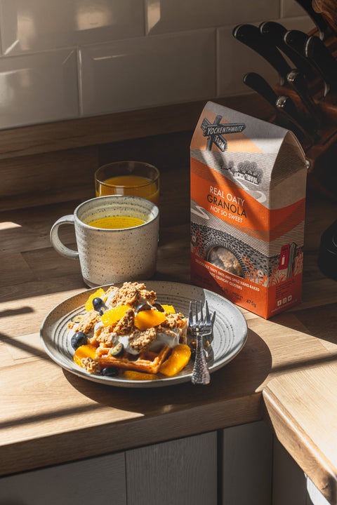 A bag of Yackenthwaite real oaty granola sits on a wooden kitchen counter next to a plate of granola with fruit, a mug of yellow drink, and an orange juice glass.