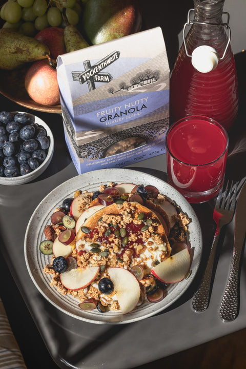 A styled breakfast scene featuring a bowl of granola topped with sliced apples, blueberries, and nuts, with a bag of Farm Fruity Nut Granola in the background alongside blueberries, a glass of red juice, and a bottle on a gray table.