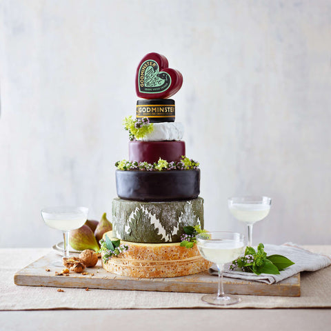 A styled cheese board featuring four stacked cheese wheels with dark red, dark brown, green, and orange rinds, topped with a heart-shaped Godminster cheese slice and garnished with herbs and grapes. Two filled coupe glasses, figs, and nuts are arranged on a wooden board against a light textured background.