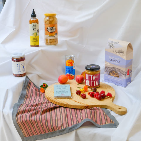 Assorted food products including jars, a box, and a cutting board with fruits on a white fabric background.