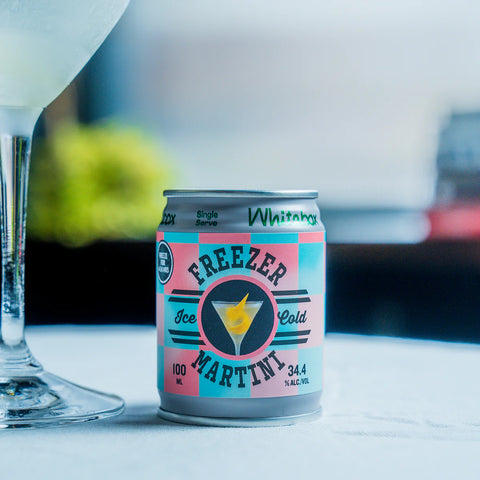A single can of Freezer Martini Ice Cold 34.4% alcohol-free cocktail mix sits on a light tabletop next to a glass of white cocktail, with a blurred background.