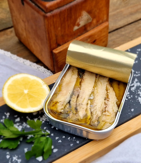 Tin of pickled fish with a gold lid on a wooden surface with a lemon and parsley.
