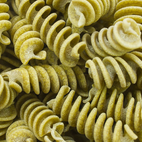 A close-up view of rotini pasta filling the entire frame, showing the spiral shape and textured surface of the dried or cooked pasta in a light olive-green color.