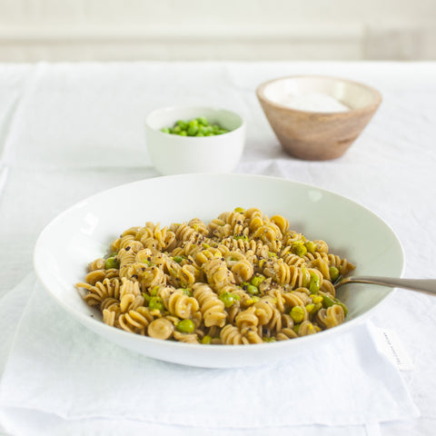 A white bowl filled with rotini pasta mixed with green peas and a light sauce, set on a white tablecloth with a spoon in the bowl and two small side dishes in the background.