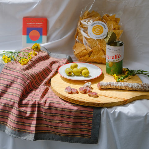 Table setting with a bag of tortilla chips, a can of Perello, and a plate of olives on a striped cloth.