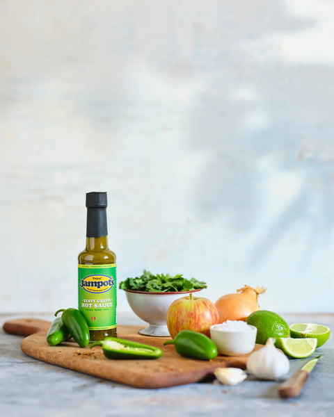 Bottle of Sambro's hot sauce on a wooden cutting board with vegetables and a bowl of salad.