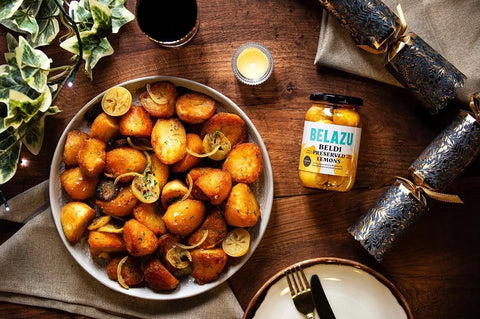 A styled tabletop scene featuring a large white bowl filled with roasted golden-brown potatoes and lemon slices, alongside a jar of Belazu Belted Lemon Pickles, decorative Christmas crackers, a candle, and a dark drink on a wooden table.