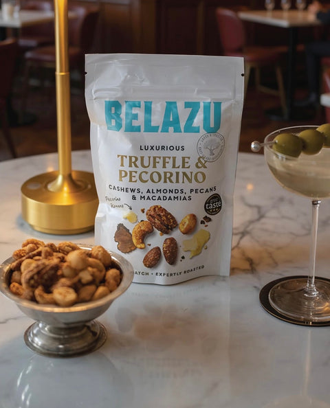 A bag of Belazu Truffle & Pecorino cashews, almonds, pecans, and macadamias sits on a white marble table, styled with a small bowl of nuts and a martini glass in a restaurant setting.