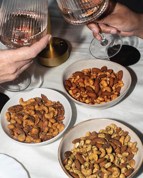 Three bowls of assorted nuts on a white tablecloth, with hands holding wine glasses in the background.