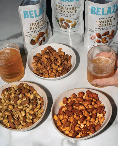 Four bags of BELAZY gourmet nuts and cashews arranged on a white marble surface with two bowls of mixed nuts and two glasses of iced coffee, with a hand holding one glass.