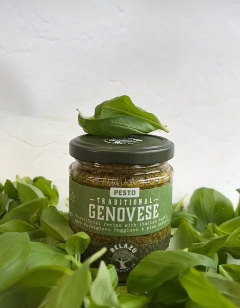 A glass jar of Belazu Traditional Genovese Pesto surrounded by fresh green basil leaves, with a single basil leaf resting on the dark green lid.
