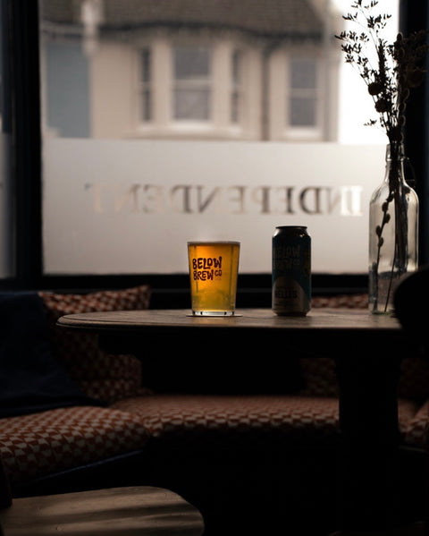 A pint of amber beer in a clear glass with 'BELOW ZERO CO.' printed in orange, next to a dark can of beer, set on a dark wooden table in a dimly lit bar or cafe interior with a window in the background.