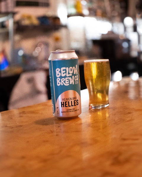 A can of Below Brew Co. 'Heaven and Hellies' non-alcoholic beer sits on a wooden bar counter next to a glass of golden beer, with a blurred bar interior in the background.
