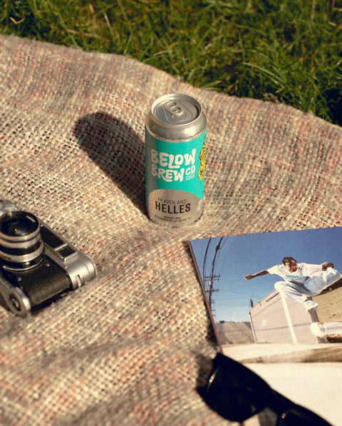 A can of Below Crew Co Helles cold brew coffee sits on a woven blanket outdoors on grass, styled with a vintage camera, sunglasses, and a printed photo.