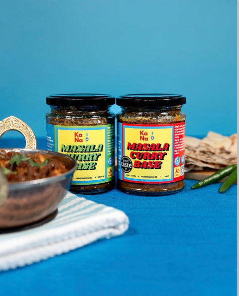 Two jars of Ka Na Masala Curry Base spices displayed on a blue tablecloth with a bowl of food and flatbread in the foreground.