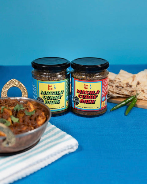 Two jars of KaNa Masala Curry Base shown next to a prepared curry dish in a bowl, flatbread, and green chilies on a blue background.