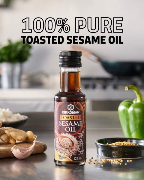 A bottle of Kikkoman Toasted Sesame Oil stands on a kitchen counter with ingredients like ginger, garlic, a green bell pepper, and bowls of sesame seeds around it. Large text at the top reads '100% PURE TOASTED SESAME OIL'.