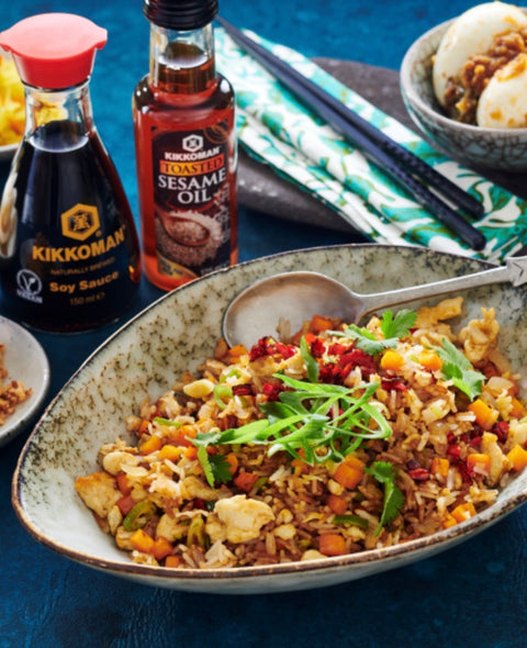 A bowl of fried rice topped with green onions and red peppers, styled with Kikkoman soy sauce, Kikkoman roasted sesame oil, chopsticks, and a small bowl of buns on a dark blue textured background.