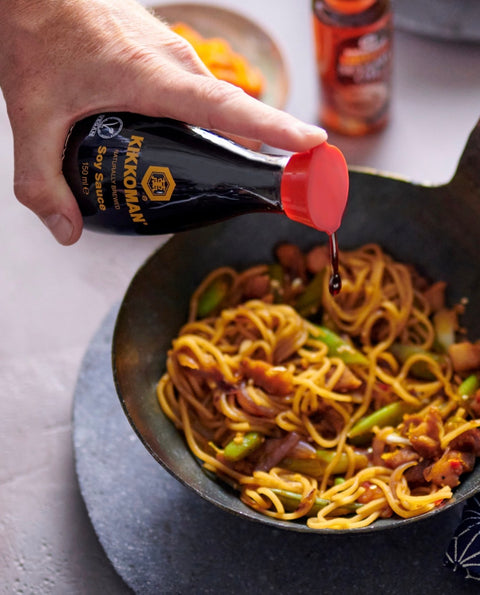 A hand pours Kikkoman soy sauce from a black bottle with a red cap into a black wok containing a stir-fried noodle dish with green vegetables and brown pieces, set on a gray surface.
