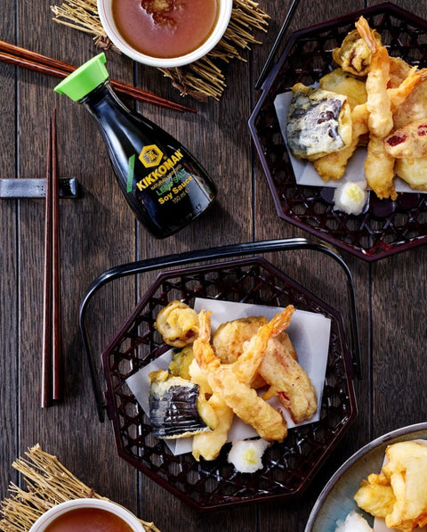 A styled overhead food scene on a dark wooden table featuring two black hexagonal baskets filled with golden fried tempura, a bottle of Kikkoman soy sauce, chopsticks, dipping sauces, and small garnish pieces.