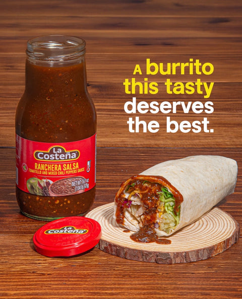 A glass jar of La costena Ranchera Salsa stands on a wooden table beside a cut burrito on a wood slice, with the red lid placed in front. Yellow and white text reads, 'A burrito this tasty deserves the best.
