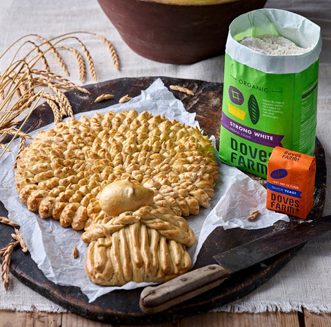 A styled product shot of baked breads and a bag of Dove Farm Strong White Flour and Dove Farm Quick Yeast on a rustic dark wooden board, with wheat stalks and a bowl in the background.