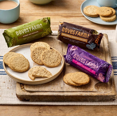 Dove Farm Ginger Oat Biscuits, Wholemeal Digestive Biscuits, and Fruity Oat biscuits are arranged on a wooden board with several biscuits on plates, set on a rustic wooden table.