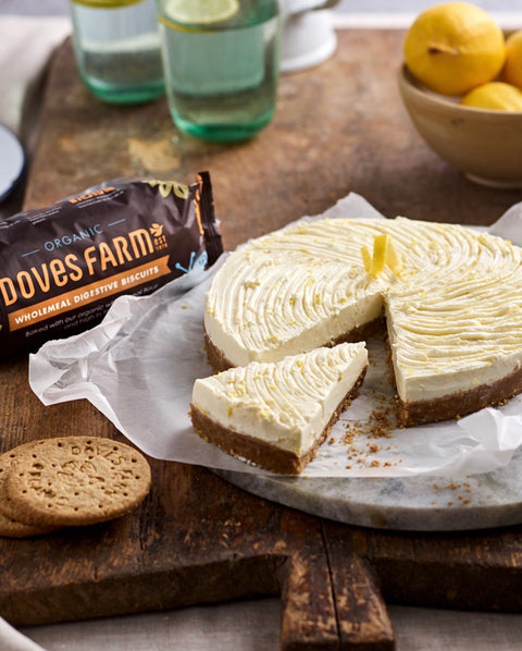 A slice of lemon cheesecake with a crumbly cookie crust sits on parchment paper on a rustic wooden board, with a package of Dove's Farm wholemeal digestive biscuits and a biscuit in the foreground and lemons in the background.