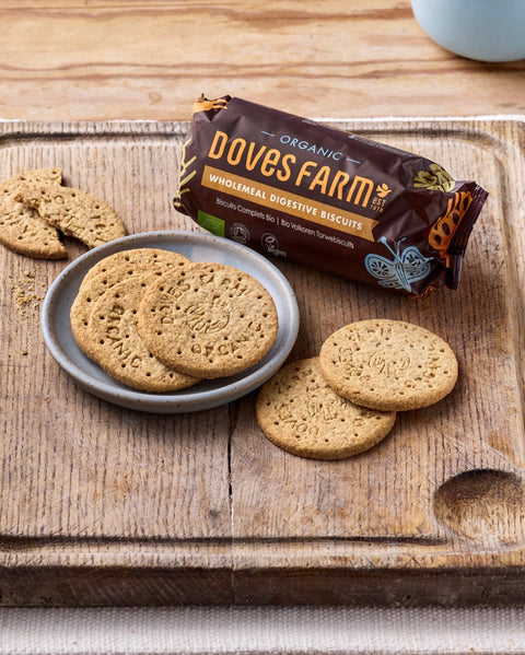 Doves Farm Organic Wholemeal Digestive Biscuits shown with several round golden-brown biscuits on a wooden board and a brown branded package.