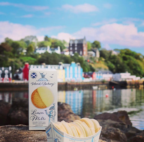 A box of Island Bakery Lemon Melt chocolate cookies stands on a rock in front of a blurred coastal town and harbor background, with a small paper tray of round cookies in front.