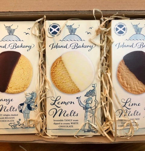 Three boxed Island Bakery Lemon Melts cookies in a cardboard tray with shredded paper filler. The packages show illustrations and photos of the cookies, with one center box displaying a tan cookie half dark chocolate and half creamy white.