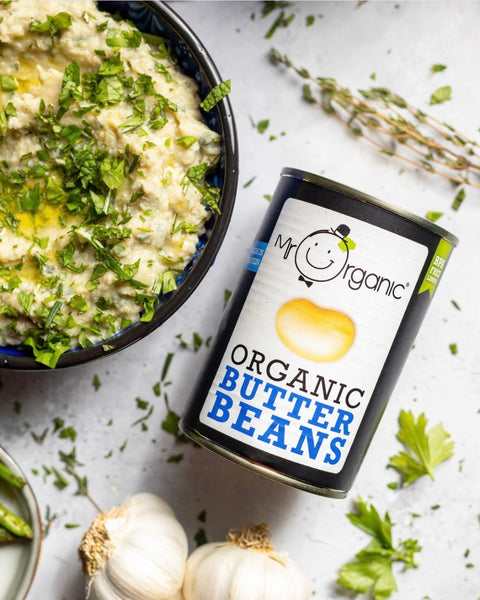 A can of My Organic Organic Butter Beans placed next to a bowl of prepared mashed beans topped with chopped parsley and oil, styled with garlic, herbs, and parsley on a light textured surface.