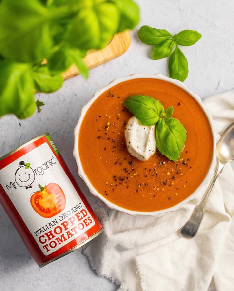 A bowl of tomato soup topped with a dollop of cream and fresh basil leaves, styled on a light surface with a can of Mr. Organic Italian Organic Chopped Tomatoes and a spoon nearby.