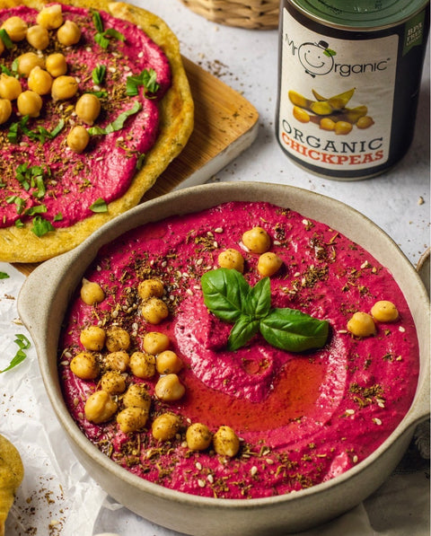 A bowl of pink chickpea soup topped with chickpeas, herbs, and a basil leaf, with a can of Organic Chickpeas in the background and a flatbread dish in the upper left.