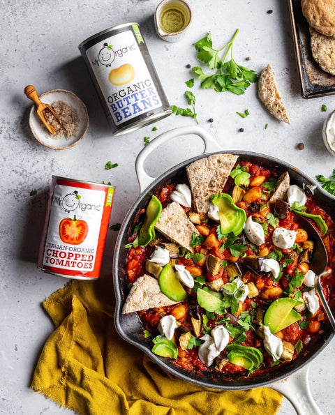 Overhead flat-lay of a prepared Mexican-style bean and tomato dish in a cast-iron skillet, with cans of organic beans and tomatoes, tortilla chips, avocado, sour cream, and herbs on a light stone surface.