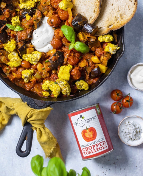Overhead flat-lay of a cast-iron skillet filled with a colorful vegetable and chickpea stew, topped with bread slices, a dollop of white sauce, and basil leaves. A can of My Organic Chopped Tomatoes is shown below the skillet on a light gray surface with small bowls of ingredients and a yellow cloth nearby.