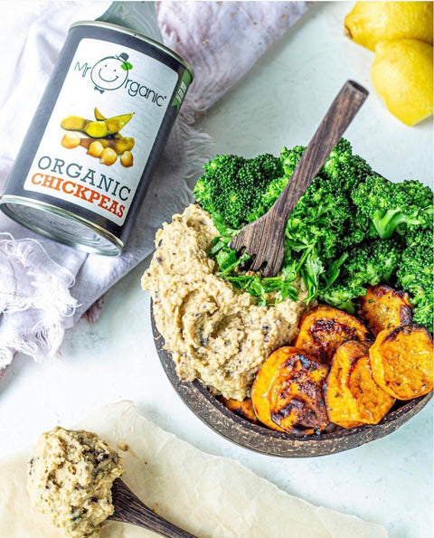 A styled food scene showing a bowl of prepared chickpea salad with broccoli, sweet potato, and hummus, alongside a can of Organic Chickpeas and a spoonful of chickpea paste on parchment paper.