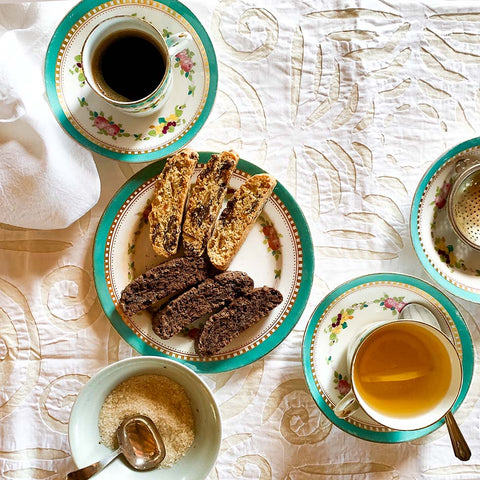 An overhead styled tea and biscuit spread on a white embroidered tablecloth. A central plate holds two types of biscuit, surrounded by teacups of black coffee and tea, a small bowl of sugar, and matching floral teacups and saucers.