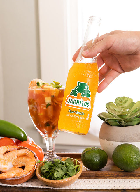 hand holding bottle of jarritos with limes, shrimp, and other foods in the background