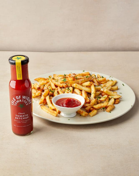 A bottle of Use of War Is Over Tomato Ketchup stands next to a plate of golden french fries with a small white dipping bowl of ketchup on a beige tabletop.