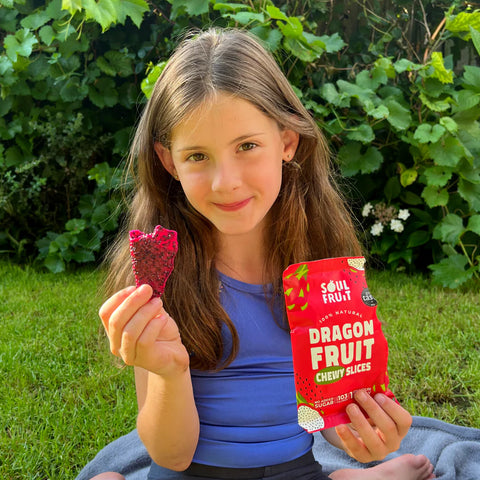 A young girl sitting outdoors on a blanket in a garden, holding a red pouch of Soul Fruit Dragon Fruit Chewy Slices in one hand and showing a piece of the fruit in the other hand.