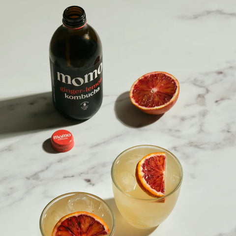 A bottle of Momo ginger lemon kombucha and its cap on a white marble surface, alongside two glasses of the pale yellow drink garnished with a blood orange slice, with a whole blood orange half nearby.