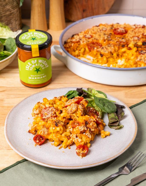 A plated serving of tomato and cheese macaroni with greens on a light gray plate, with a jar of Makhani's 'Taste of India' tomato sauce and a baked casserole dish in the background on a wooden table.