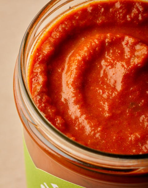 A close-up shot of a glass jar filled with thick, glossy red tomato sauce, showing the rich texture of the puree inside.