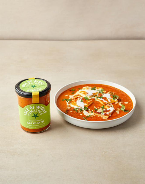 A glass jar of Taste of Wigan Makhni chutney next to a white bowl filled with orange-red curry and topped with cream, garnished with herbs, on a beige surface.