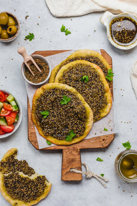 Three round flatbreads topped with a dark green herb and sesame spread, arranged on a wooden cutting board with parchment paper. The board is styled on a light gray surface with bowls of olives, sliced tomatoes and cucumbers, a small pitcher of dressing, and a broken piece of flatbread in the foreground.